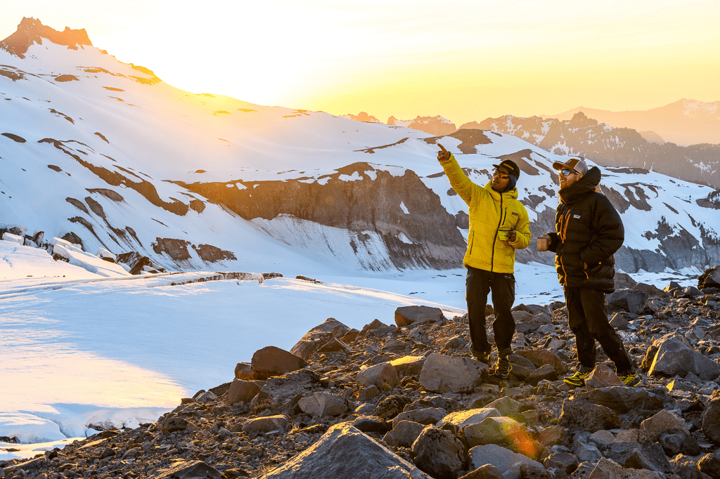 Two friends standing on a rocky outcropping in a snowy mountain range at sunrise.