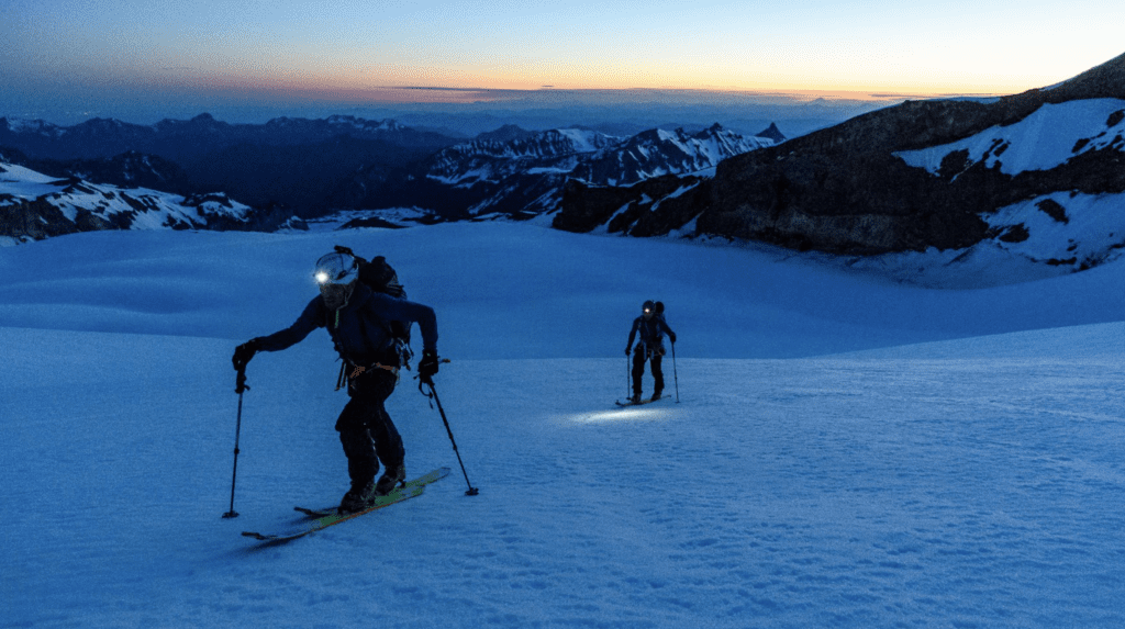 Two skiers crossing flat terrain at sunrise with headlamps on.